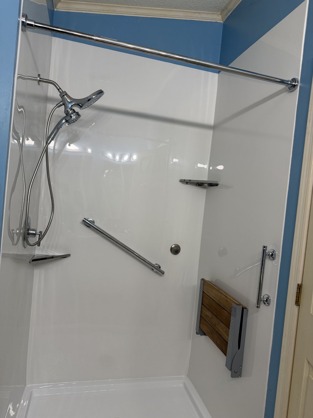 White shower with wall-mounted grab bars, handheld showerhead, corner shelves, and a folding seat
